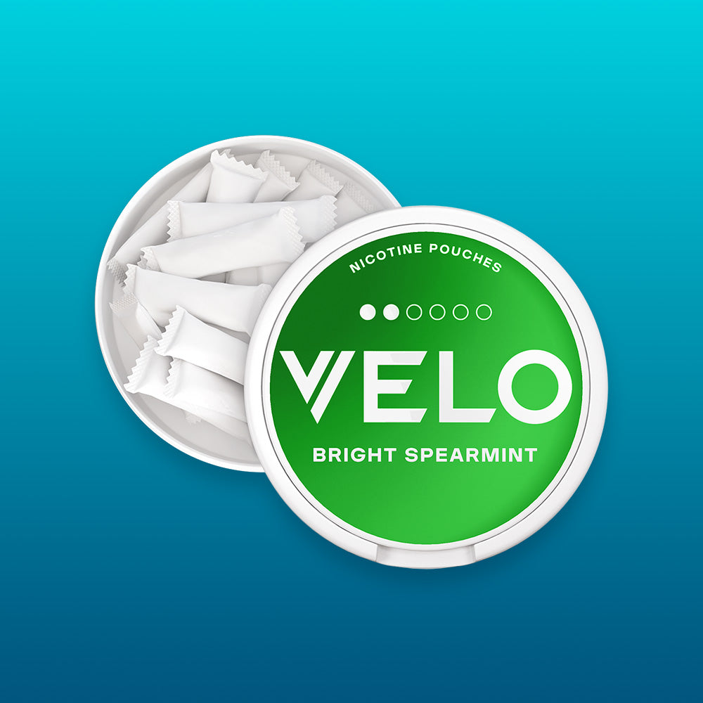 Velo Nicotine Pouches |  A round container labeled VELO Bright Spearmint holds several white nicotine pouches, with the lid partially open, displayed on a blue gradient background. The Velo pouches are neatly arranged inside, emphasizing their convenient and modern design.