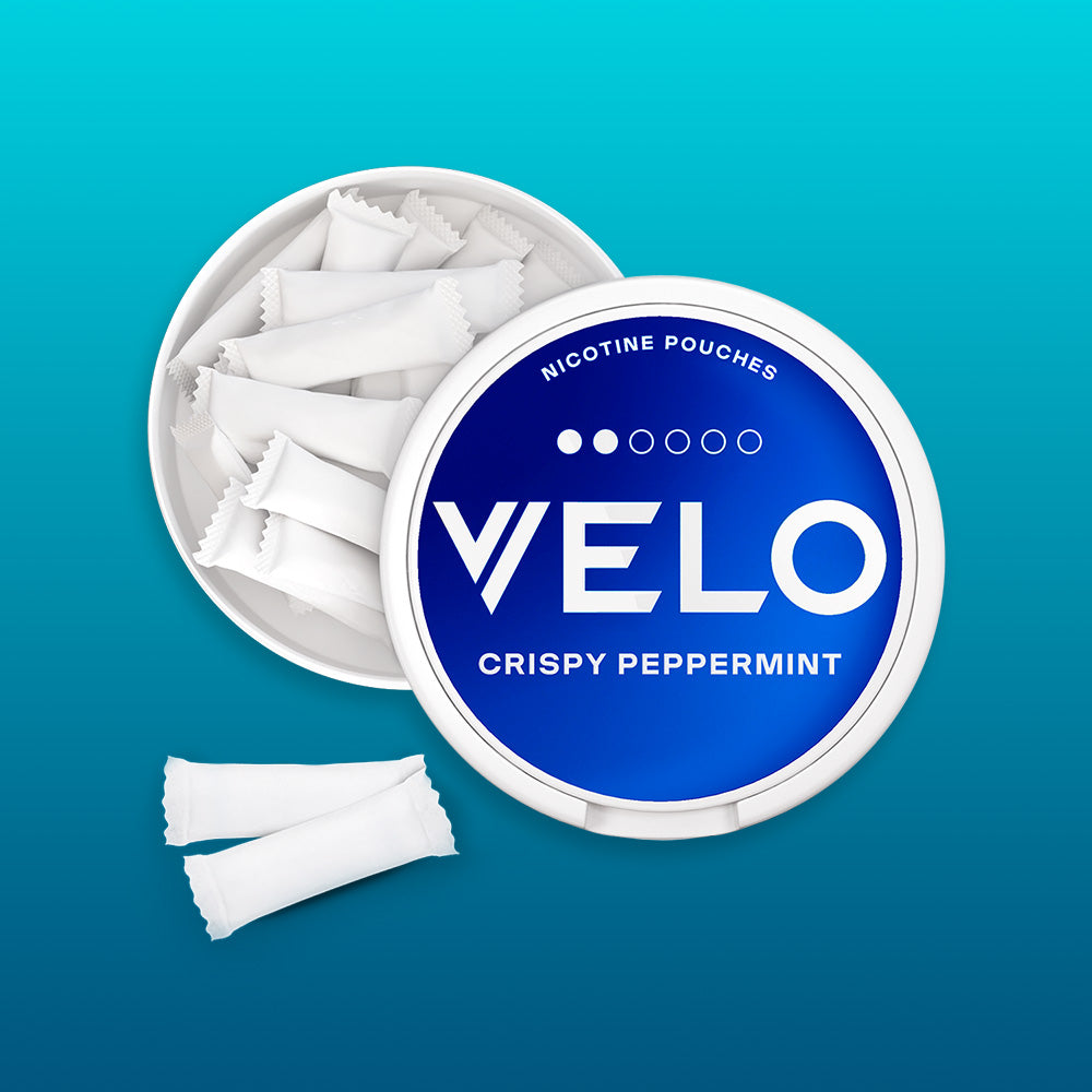 Velo Nicotine Pouches |  A round, blue and white container labeled “VELO CRISPY PEPPERMINT NICOTINE POUCHES” sits against a blue gradient background, with several Velo nicotine pouches neatly arranged inside and a few loose pouches displayed outside.