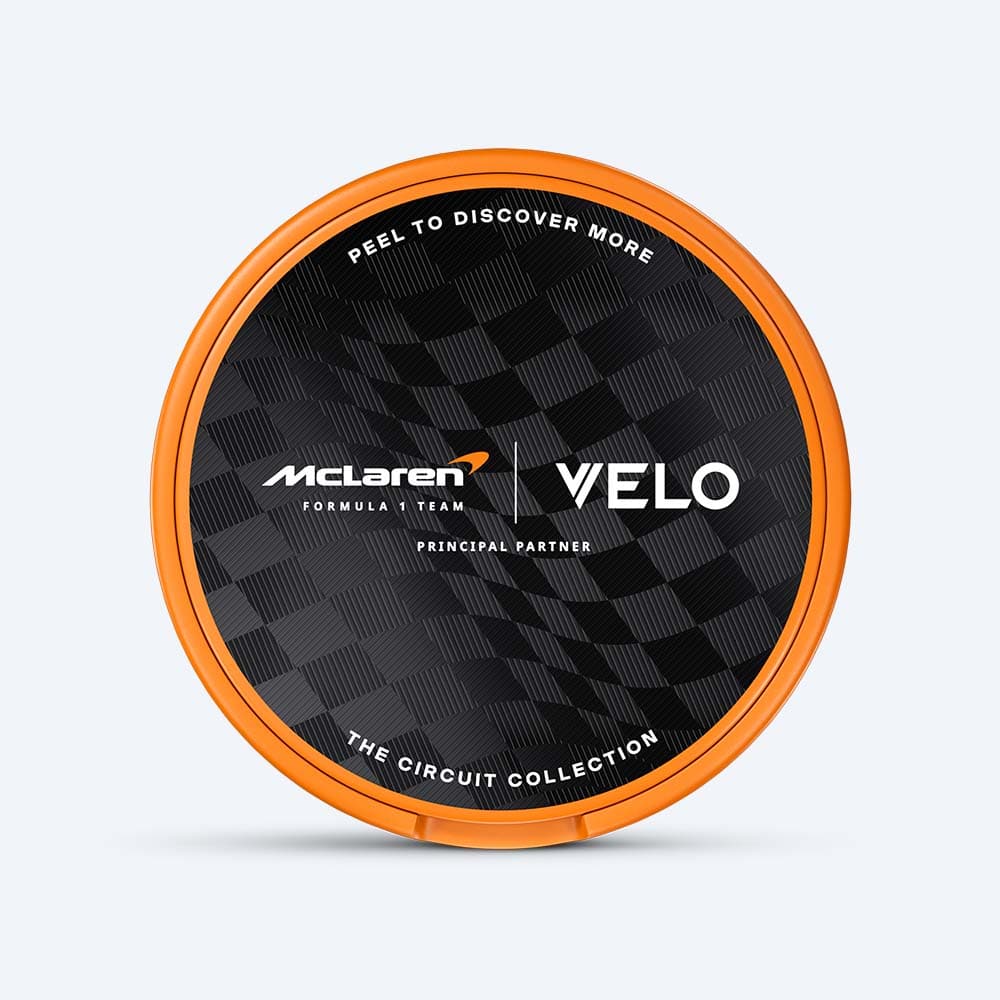 Velo Nicotine Pouches |  Round orange tin with a black checkered lid, showcasing the VELO and McLaren Formula 1 Team logos in white, along with the phrases "Principal Partner" and "The Circuit Collection." The vibrant design from the VELO Limited Edition Circuit Collection, inspired by McLaren Racing, hints at tropical flair—capturing the papaya flavour’s energy in a sleek, collectible package.