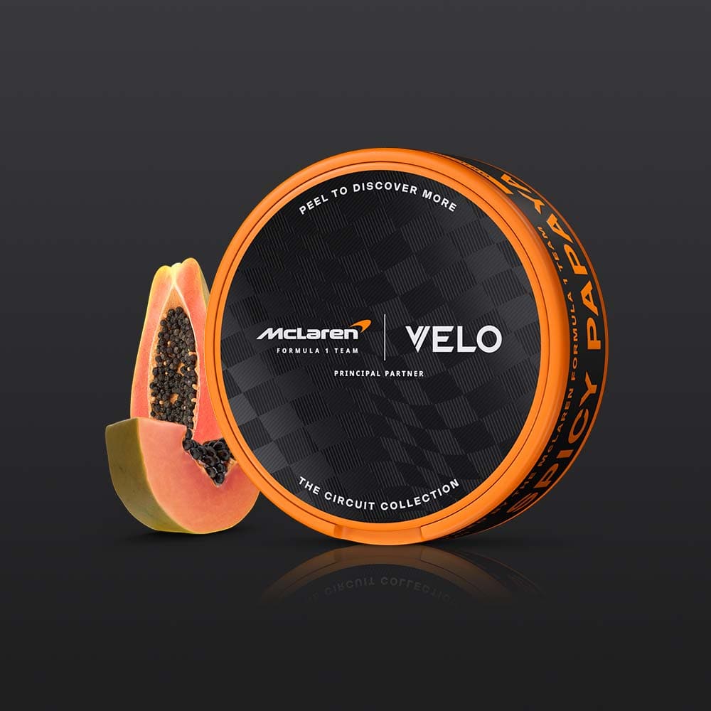 Velo Nicotine Pouches |  A round orange can of VELO Limited Edition Circuit Collection Design Inspired by McLaren Racing sits beside a halved papaya on a dark background. The can features VELO branding with references to “SPICY PAPAYA” and “THE CIRCUIT COLLECTION,” emphasizing its tropical papaya flavour.