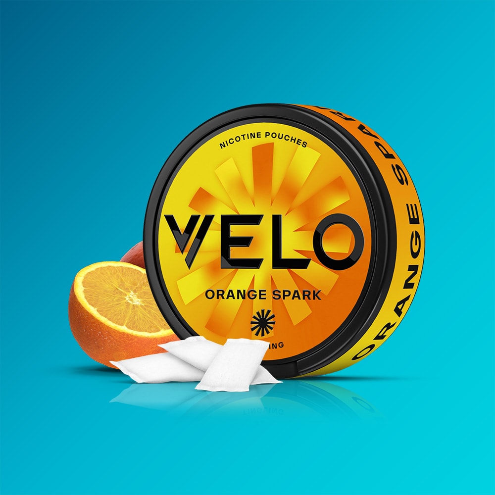 Velo Nicotine Pouches |  A round tin of VELO Orange Spark nicotine pouches rests on a teal background. The yellow and black tin features a sunburst design, evoking the vibrant citrus flavor within. Beside the tin are two white nicotine pouches and half an orange, inviting you to experience the zesty essence of Orange Spark with every use.