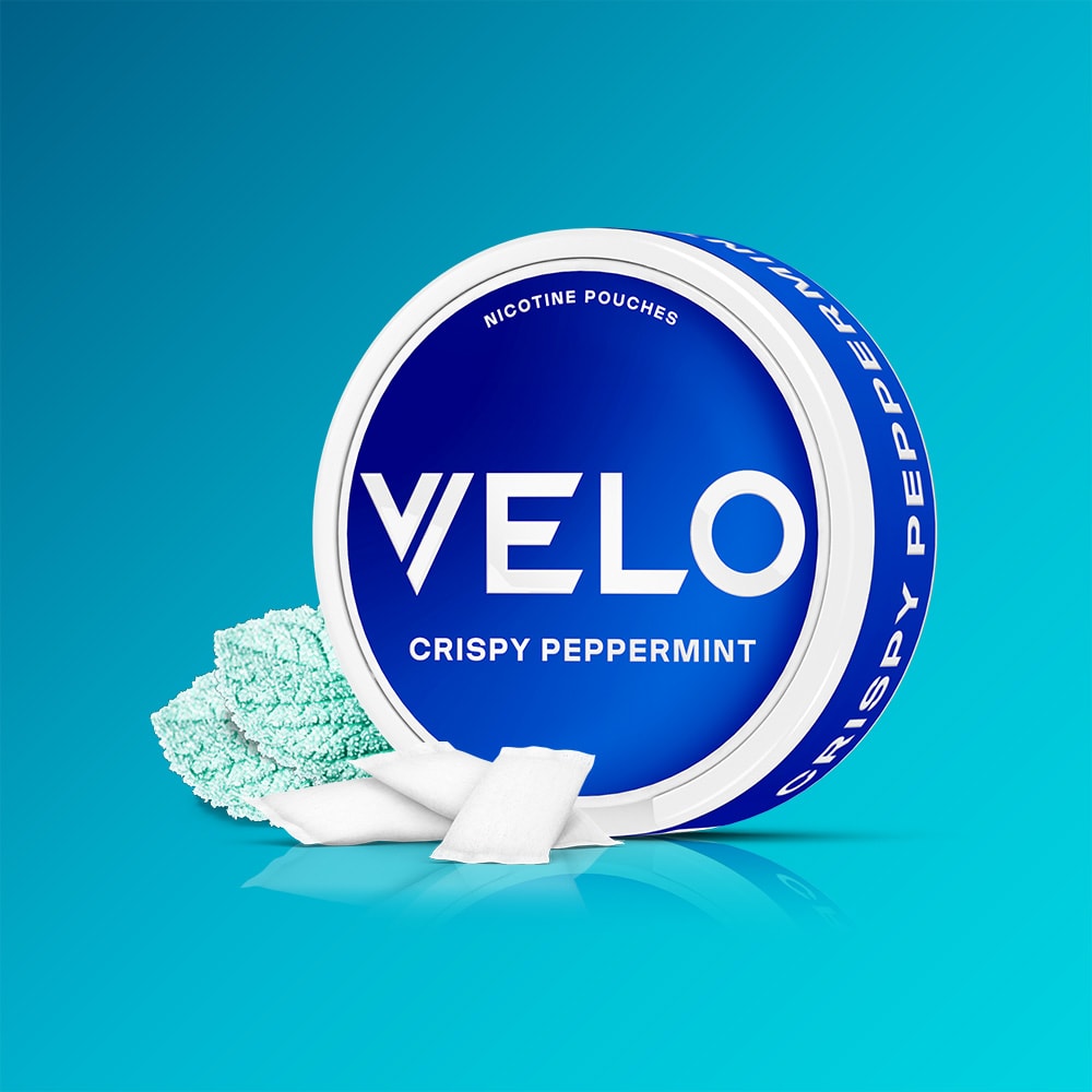 Velo Nicotine Pouches |  A circular blue VELO Crispy Peppermint container rests on a blue surface, accompanied by several white nicotine pouches and fresh mint leaves, emphasizing the peppermint flavor and signature cooling aftertaste.