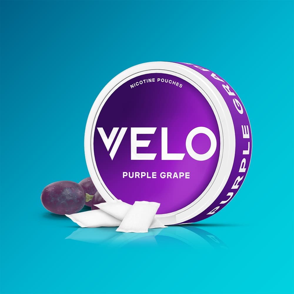 Velo Nicotine Pouches |  A round container of VELO Purple Grape nicotine pouches with a purple label is displayed on a blue background, accompanied by two red grapes and several white pouches next to it.