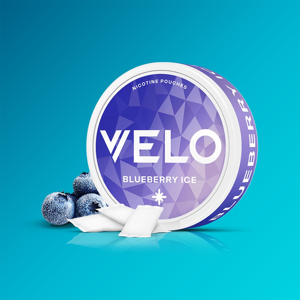 Velo Nicotine Pouches |  A round container of VELO Blueberry Ice sits on a blue surface, accompanied by frozen blueberries and several white nicotine pouches. The background features a gradient blue hue, evoking the cool sensation and fruity tartness of blueberry in every pouch.