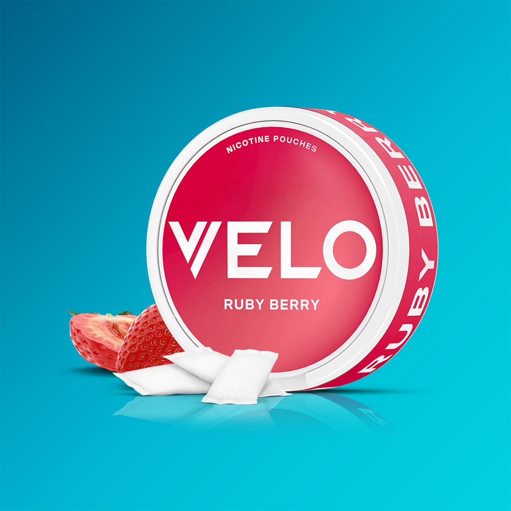 Velo Nicotine Pouches |  A round red VELO Ruby Berry container featuring nicotine pouches sits against a blue background, accompanied by a halved strawberry that highlights the fresh blend’s acidity, with white pouches displayed in front.