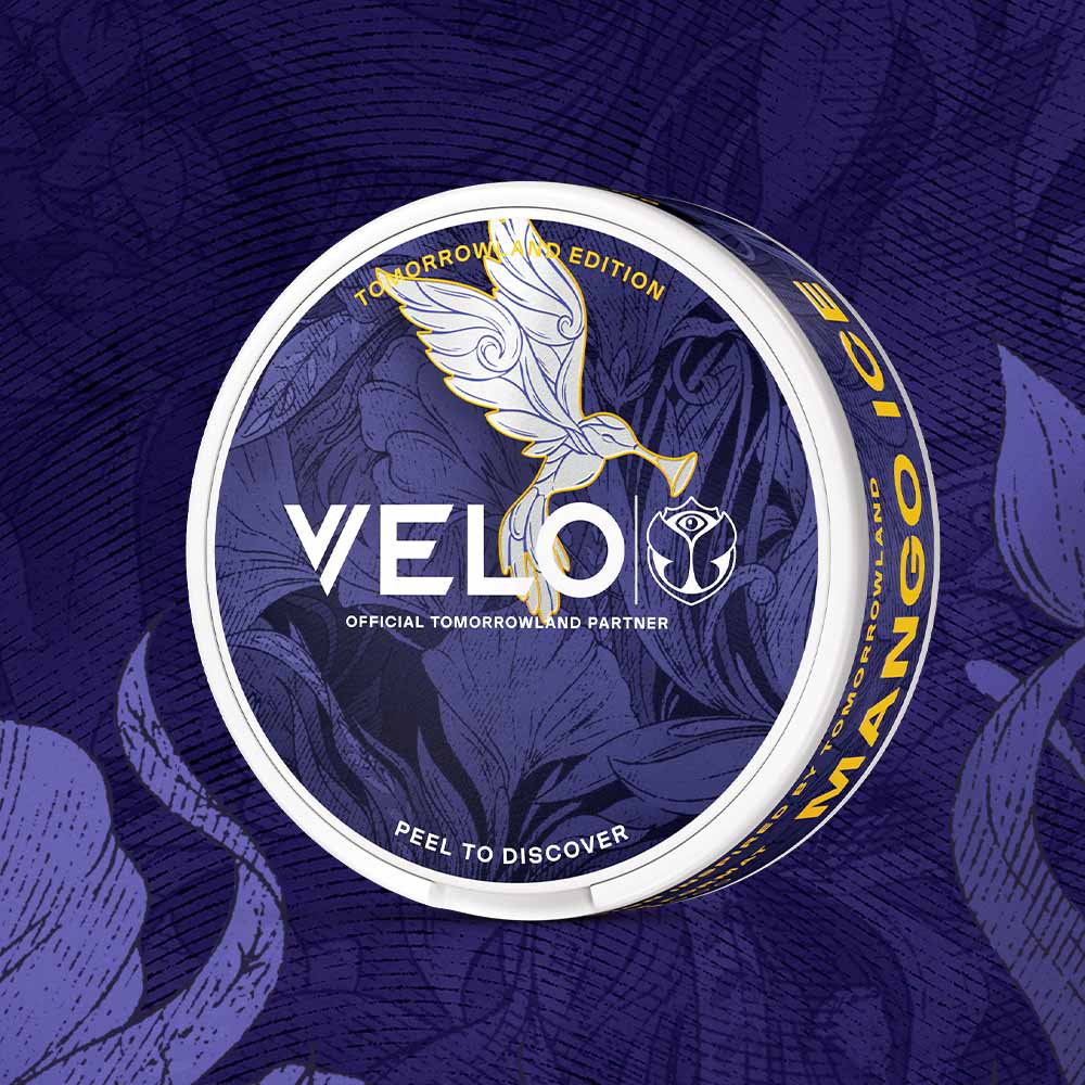 Velo Nicotine Pouches |  A round container labeled VELO Tomorrowland Limited Edition Mango Ice features a white dove and floral designs set against a purple background. The text includes “Tomorrowland Edition” and “Peel to Discover,” emphasizing this exclusive release of the refreshing VELO Mango Ice flavor.