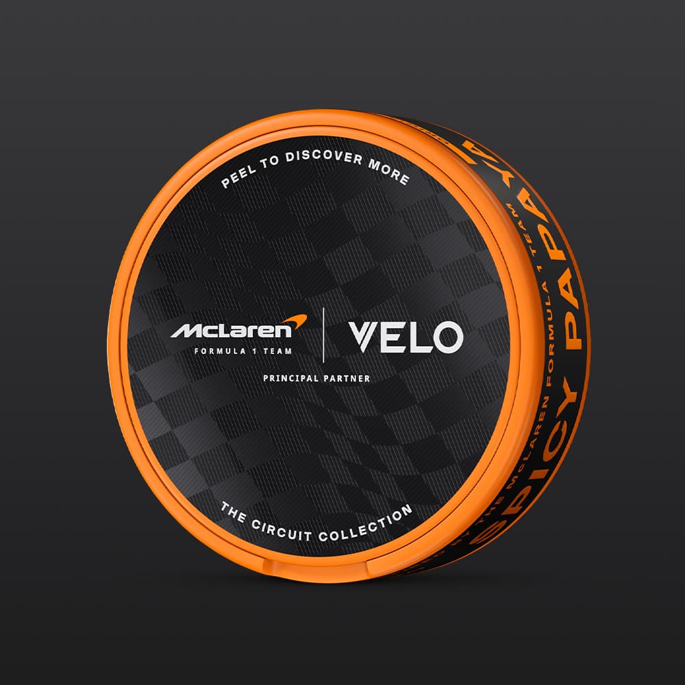 Velo Nicotine Pouches |  A round black and orange can featuring VELO branding and the McLaren Formula 1 Team, labeled as the VELO Limited Edition Circuit Collection Design Inspired by McLaren Racing, with a creamy papaya flavour and a "Peel to Discover More" message, displayed against a dark background.