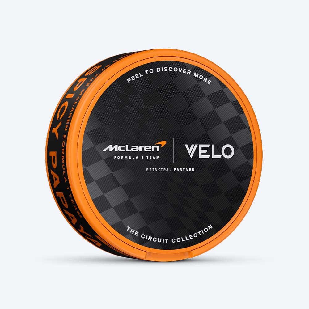 Velo Nicotine Pouches |  A round orange and black tin featuring a checkered pattern showcases the VELO Limited Edition Circuit Collection Design Inspired by McLaren Racing. The lid displays McLaren Formula 1 Team, VELO, and The Circuit Collection branding, while the rim highlights "Spicy Papaya"—suggesting a bold papaya flavor inside. Instructions invite users to peel open the tin to discover more of this tropical experience.