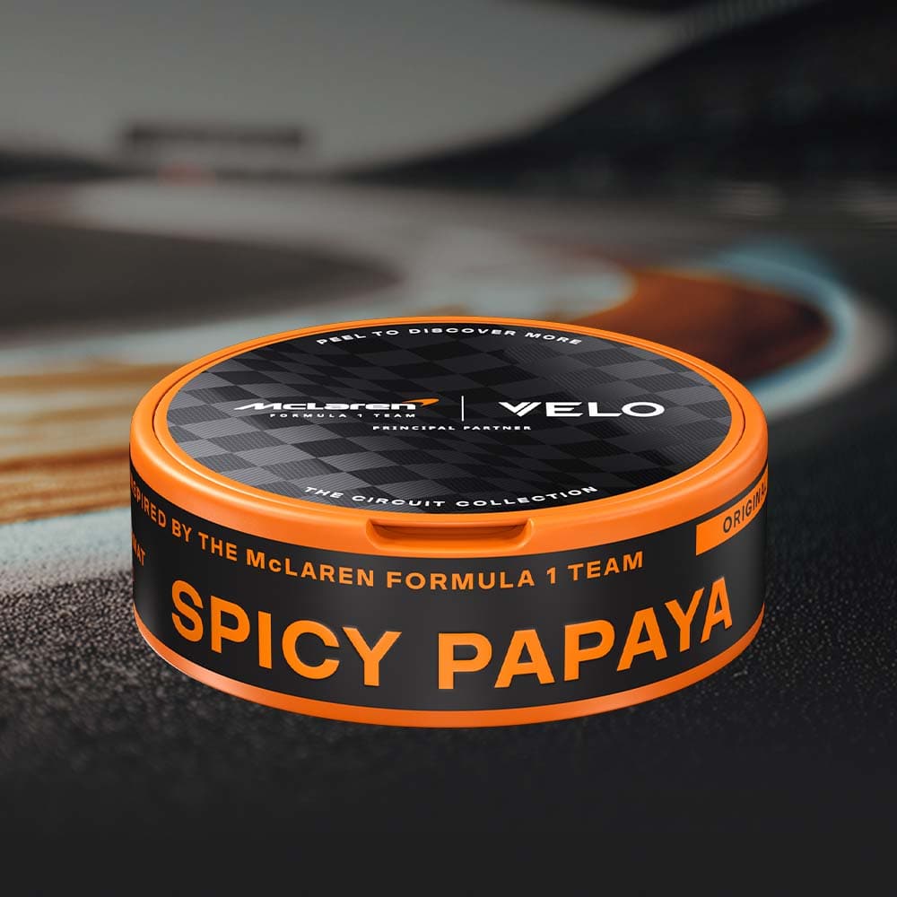 Velo Nicotine Pouches |  A round, orange and black container from VELO, featuring the Limited Edition Circuit Collection Design inspired by McLaren Racing, is displayed against a blurred racetrack background. Showcasing a creamy, tropical papaya flavour, this special edition stands out both on and off the track.