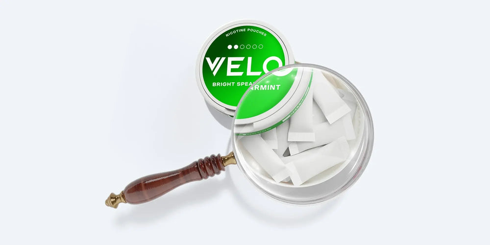 Velo Nicotine Pouches |  A tin of VELO Bright Spearmint nicotine pouches sits open, showing white pouches inside. A magnifying glass is placed over the tin, enlarging part of the Velo pouches and their crisp contents.